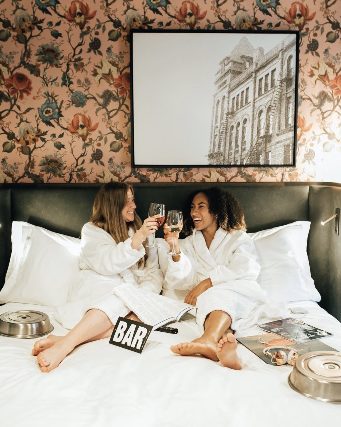 Two friends in bathrobes toasting wine glasses in a Broadview Hotel room beneath a framed drawing of the hotel